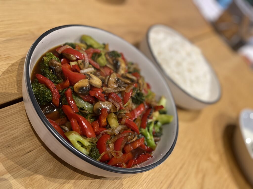 Two white bowls placed on wooden table, one filled with white rice in background, the other filled with various cooked vegetables