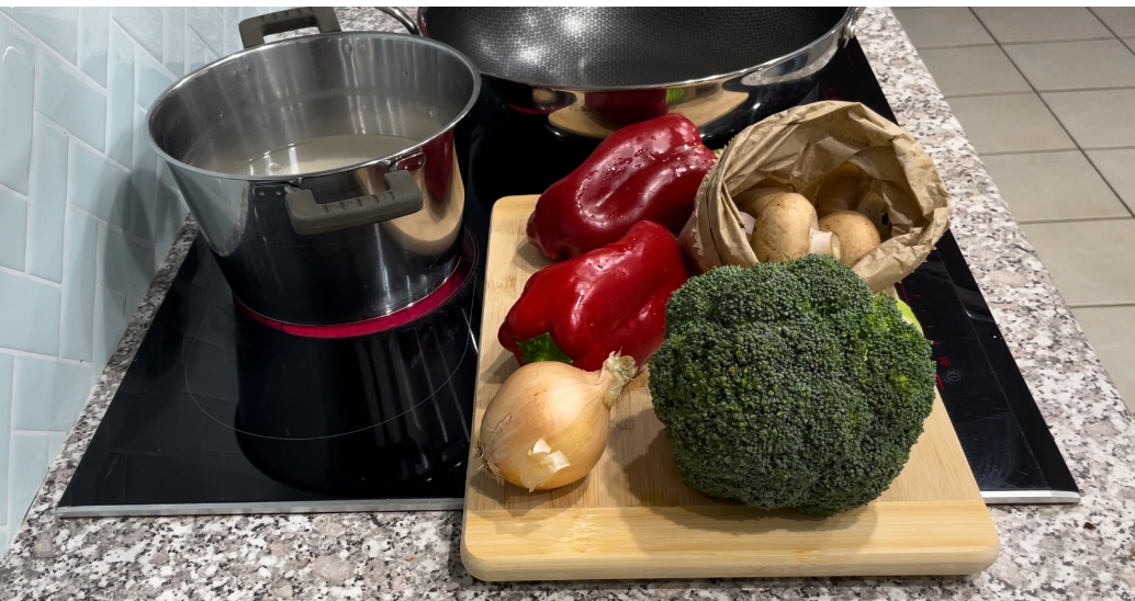 Black stove top with wooden chopping board, filled with broccoli, onion, capsicum and paper bag of brown mushrooms with silver pot and wok placed on heat