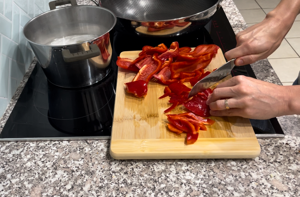 Woman chopping red capsicum on wooden chopping board placed on black stovetop. Silver pot and wok placed on heat