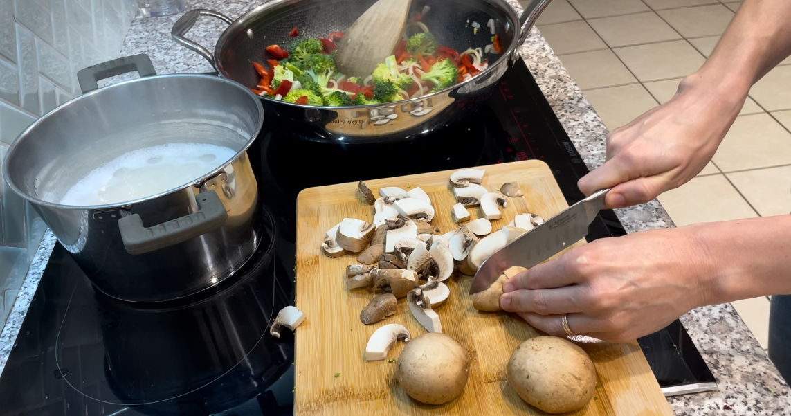 Woman slicing brown mushrooms on wooden chopping board placed on black stovetop. A pot of rice and wok filled with vegetables are placed on heat beside her.