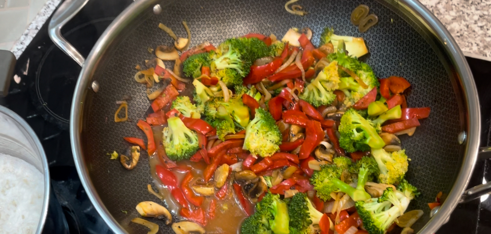 Wok on black stovetop filled with broccoli, red capsicum and onion