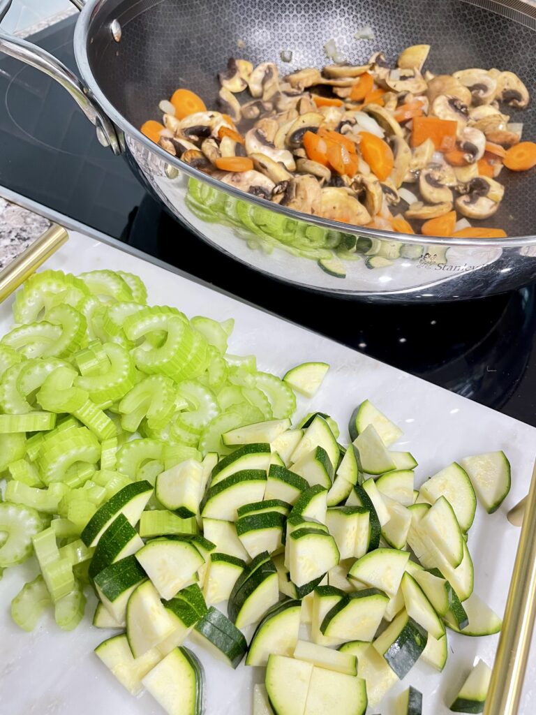 Mushrooms and carrots in silver wok on stove next to white tray with sliced zucchini and celery