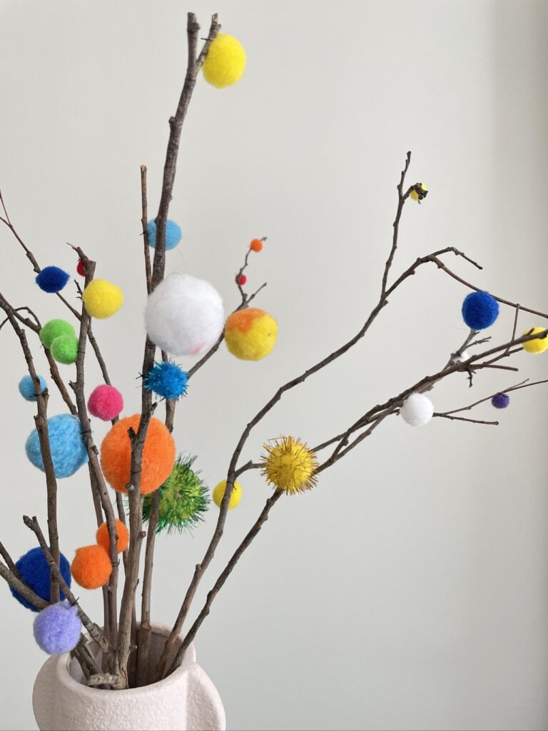 Blush vase with pom pom decorated twigs against white background