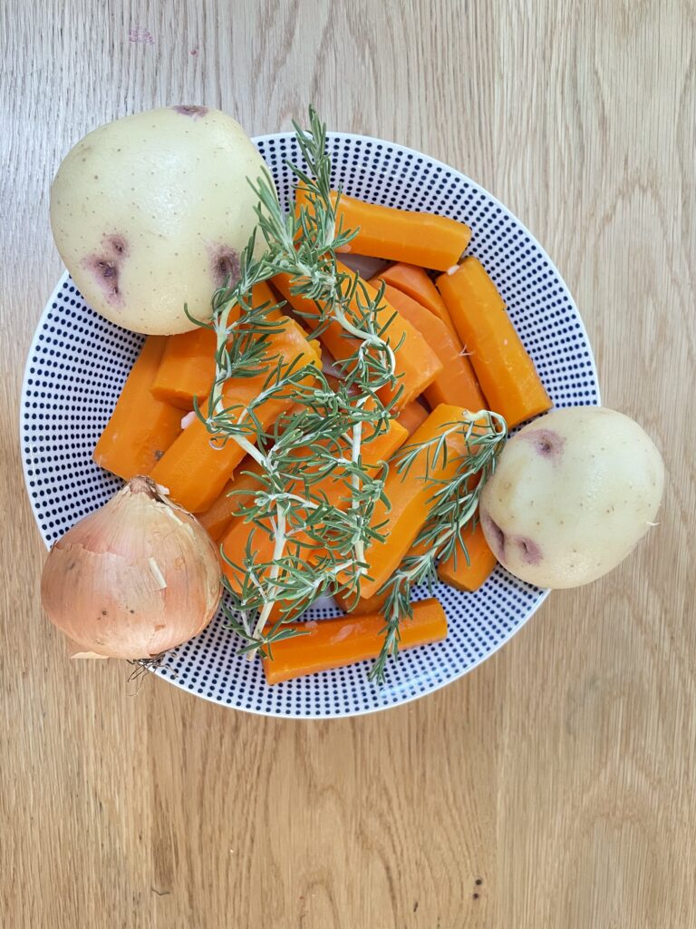 Bowl on wooden table with carrots, potatoes, rosemary and onion