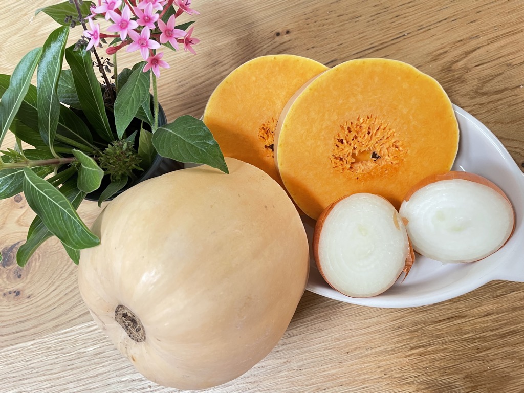 White bowl filled with sliced butternut squash and half onions Next to half butternut squash and vase of flowers