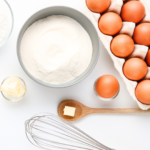 Carton of eggs, bowl of flour and whisk on white backdrop