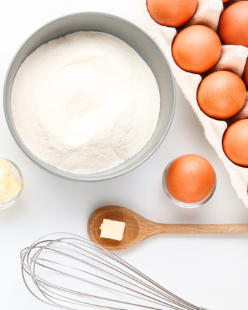 Carton of eggs, bowl of flour and whisk on white backdrop