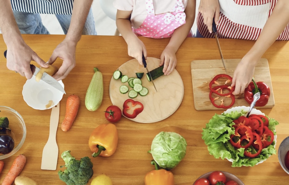 Three people in aprons on kitchen bench slicing vegetables