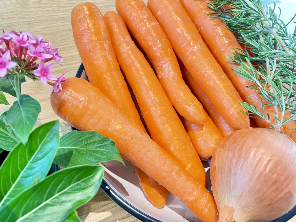 Bowl filled with fresh carrots, an onion and rosemary sprigs next to flowers