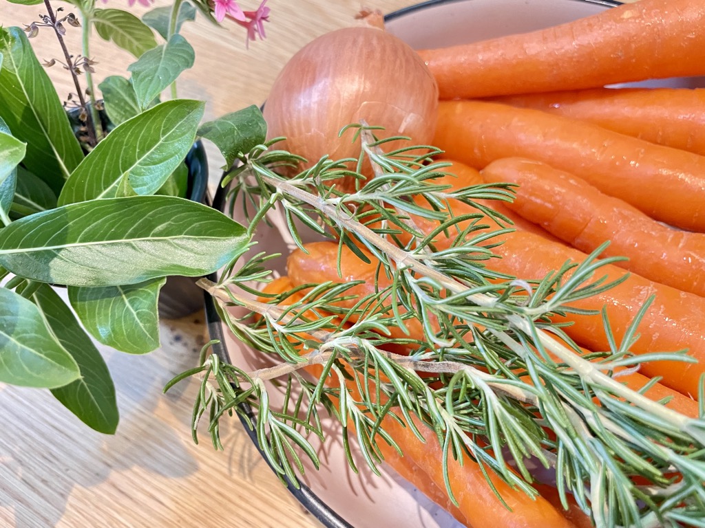 Raw Carrot sticks, onion and rosemary in bowl next to vase on wooden table