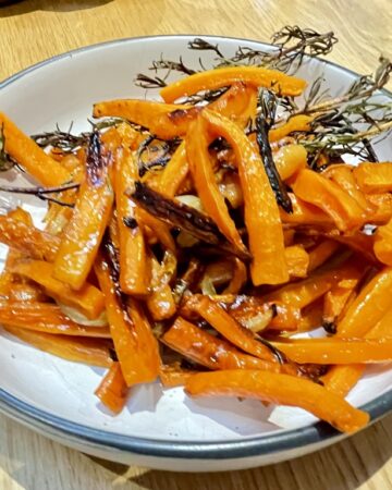 White bowl with black rim, filled with roasted carrots and rosemary On wooden table