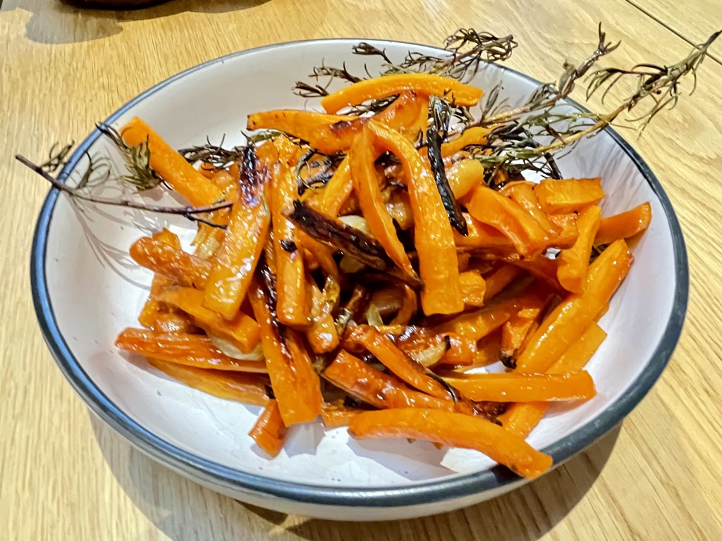 White bowl with black rim, filled with roasted carrots and rosemary On wooden table