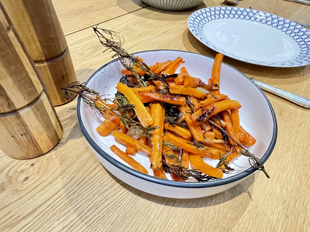 Wooden table set with plate, salt and pepper grinder and bowl with roasted carrots and rosemary