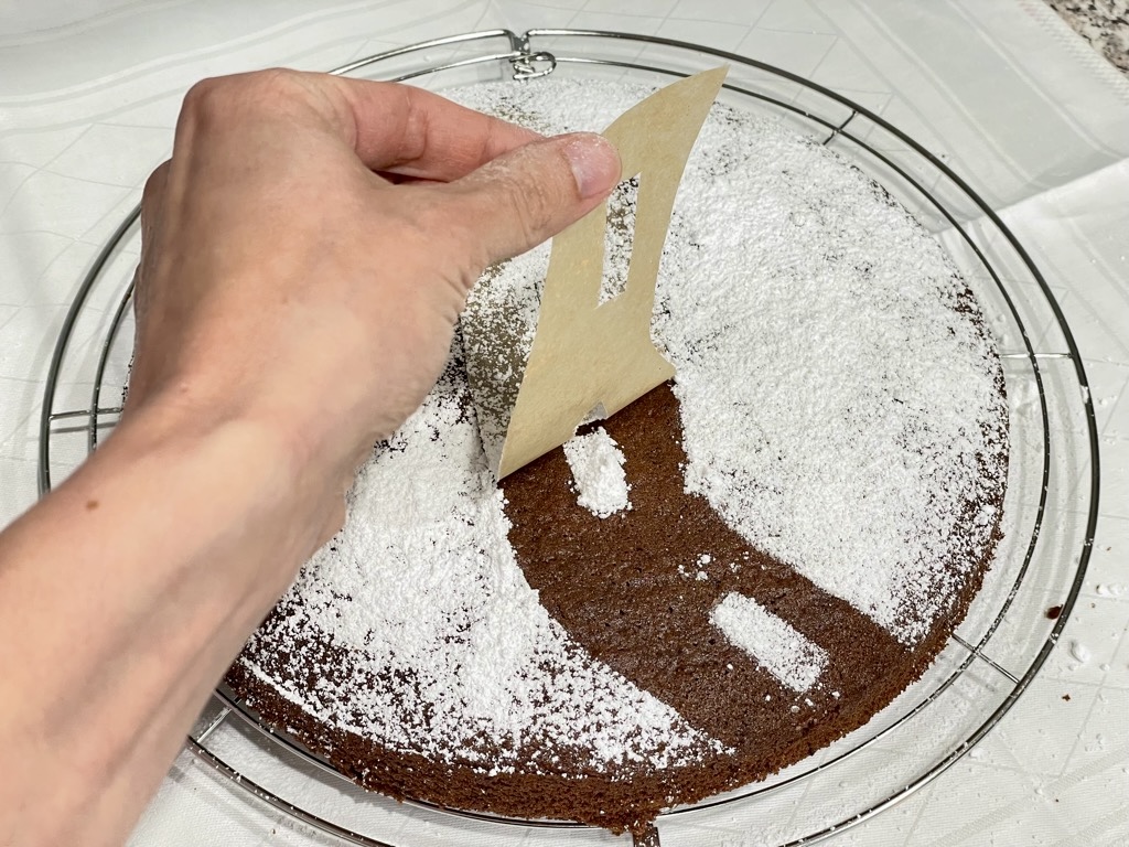 Hand lifting stencil off cake covered in powdered sugar