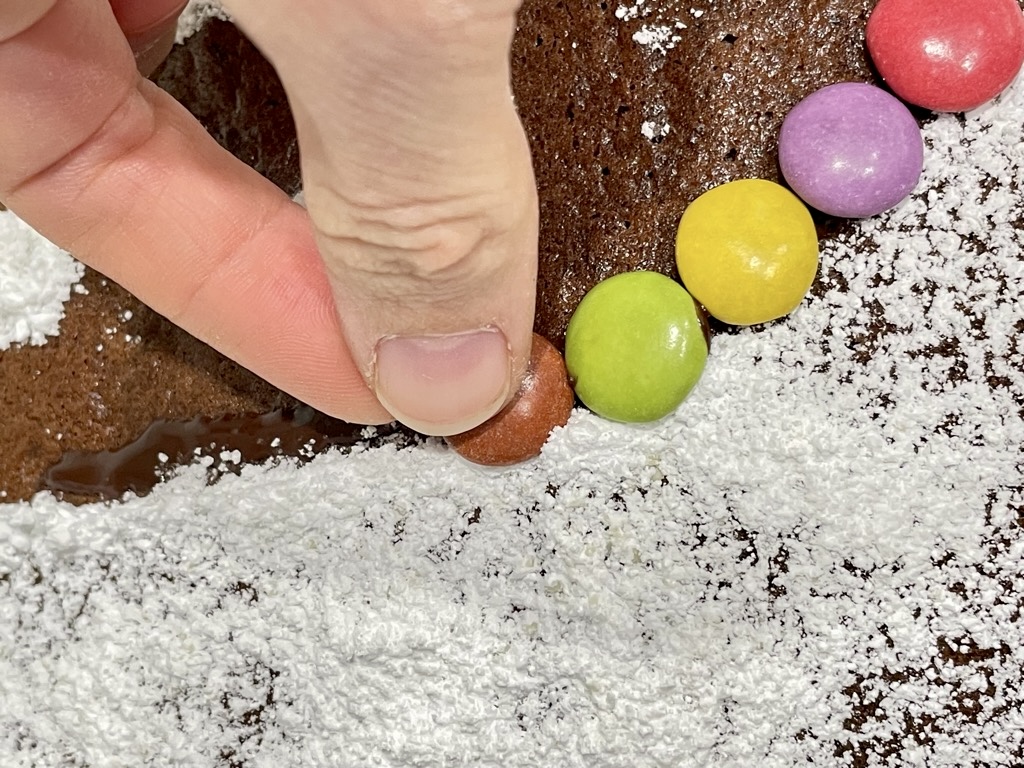 Hand placing smarties on powdered sugar and chocolate on cake