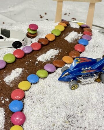 cake decorated as road design made from smarties and powdered sugar with two hot wheels cars parked on either side of the road