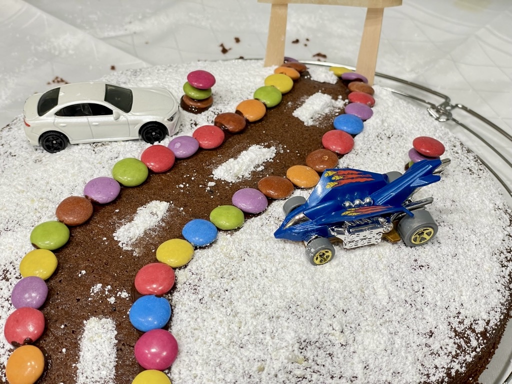 cake decorated as road design made from smarties and powdered sugar with two hot wheels cars parked on either side of the road