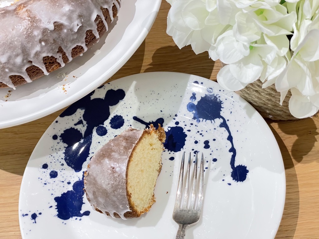 A slice of rustic homemade lemon drizzle cake on white plate with blue speckles