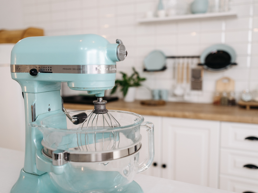 Light blue kitchen aid stand mixer with glass bowl in white kitchen with wooden bench