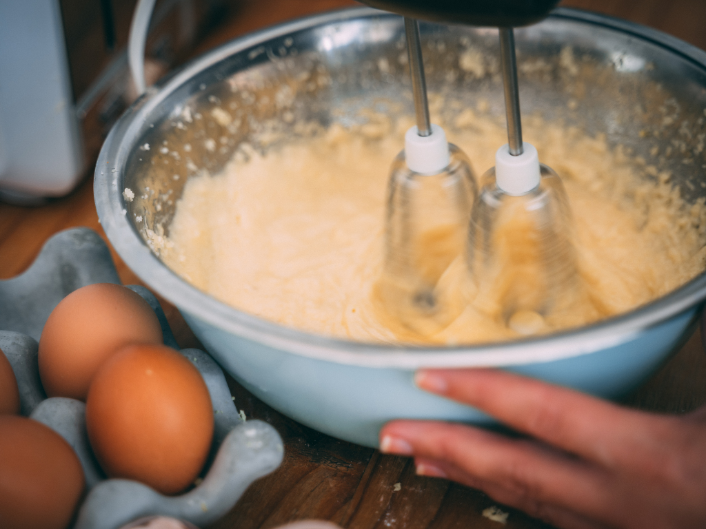 Hand holding silver mixing bowl with whisk mixing dough aside a carton of eggs