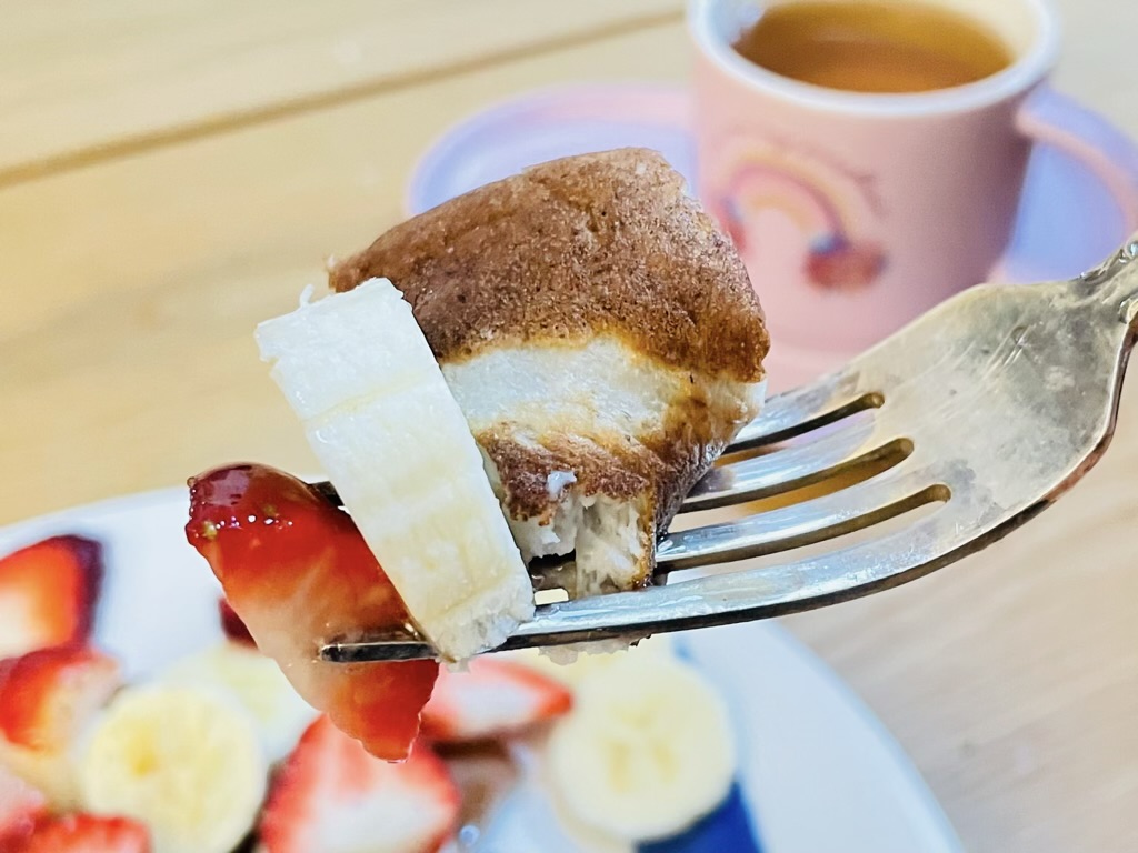 Fork holding slice of banana and strawberry with piece of pancake. Cup of tea in background