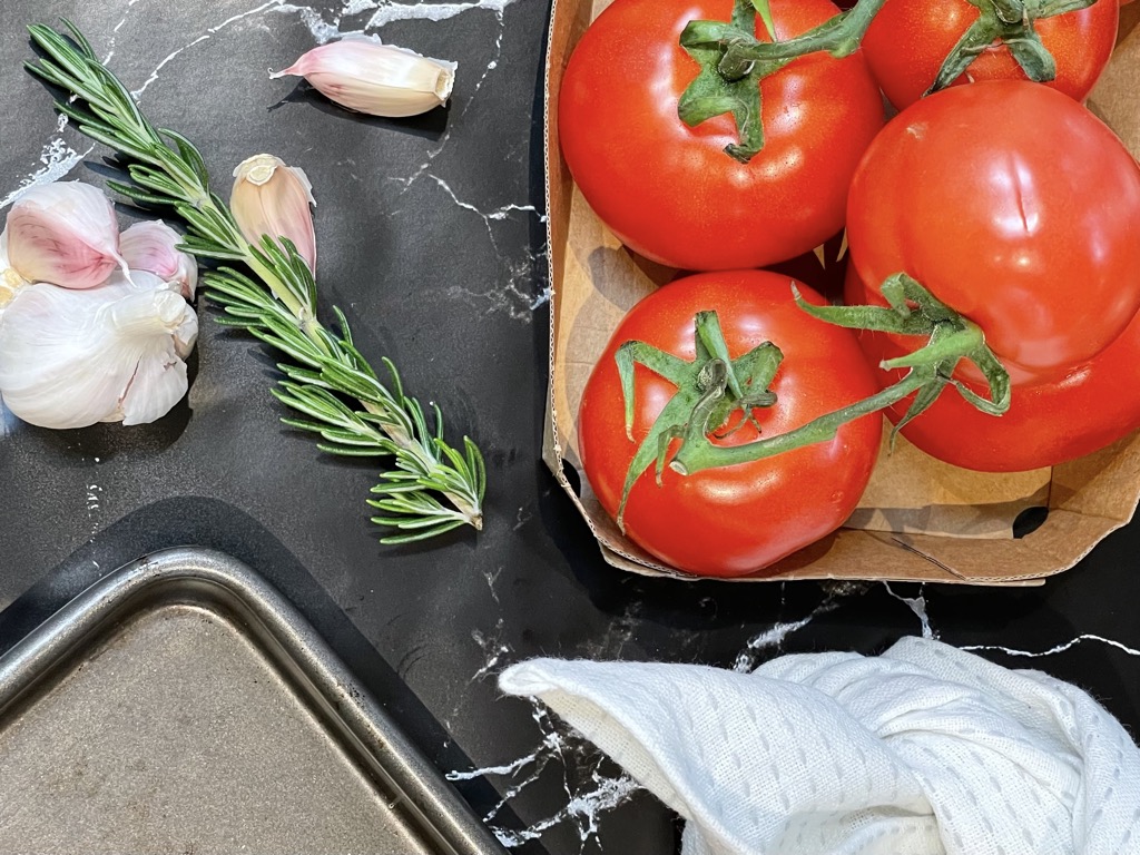 Paper tray of truss tomatoes, tied white dish cloth, garlic and rosemary and baking tray on black marble bench