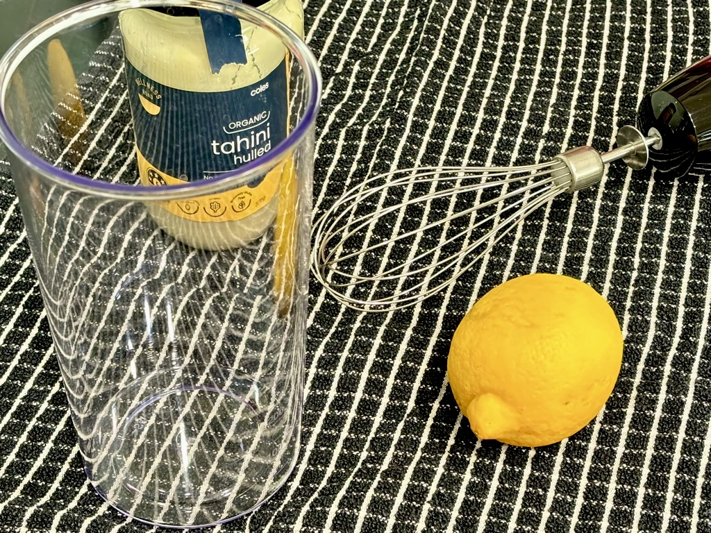 Black tea towel topped with whisk, lemon, jar of organic tahini and clear mixing jar