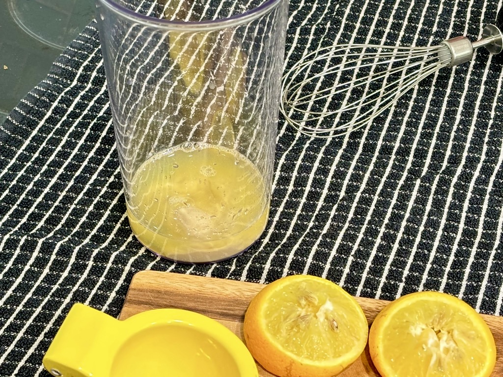 Black checkered kitchen towel topped with hand whisk, clear jar with lemon juice, squeezed lemons and juicer on wooden board