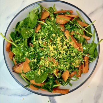 White bowl on marble surface, filled with roasted carrots and spinach, topped with lemon zest