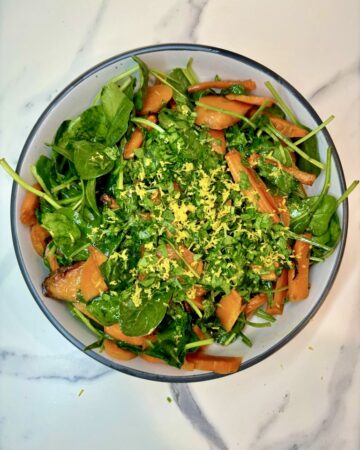 White bowl on marble surface, filled with roasted carrots and spinach, topped with lemon zest