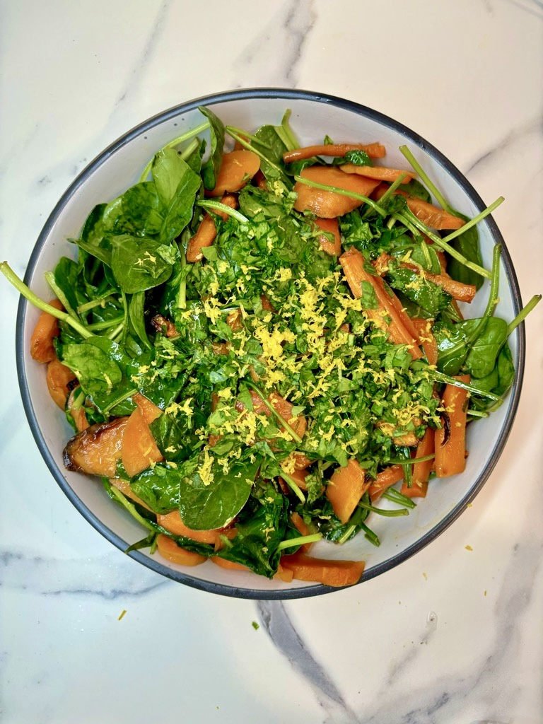 White bowl on marble surface, filled with roasted carrots and spinach, topped with lemon zest