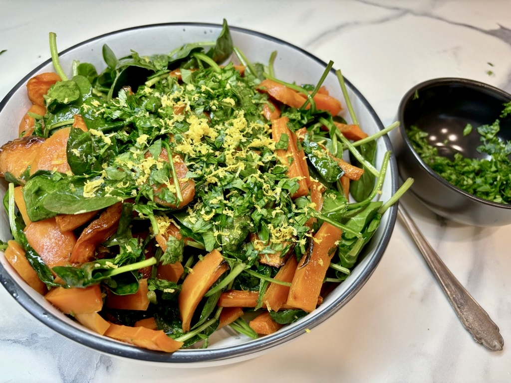 White bowl on marble surface, filled with roasted carrots and spinach, topped with lemon zest, next to black bowl of greens