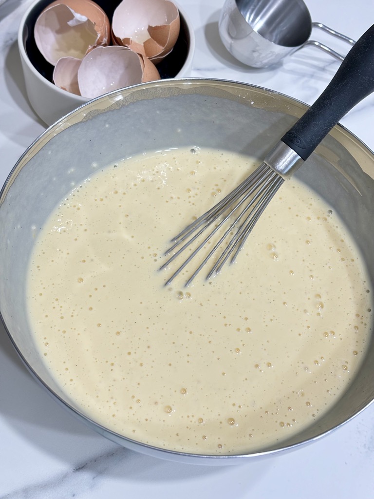 Silver bowl of batter next to pile of egg shells and measuring cup