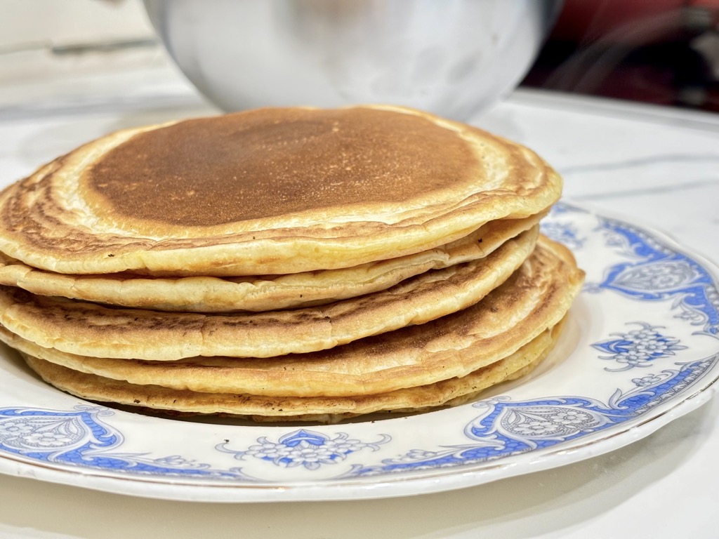 Stack of pancakes on blue and white plate in front of silver bowl