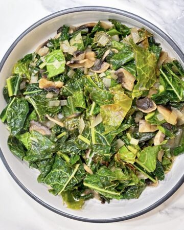 White dish filled with cooked kale and mushrooms on marble surface