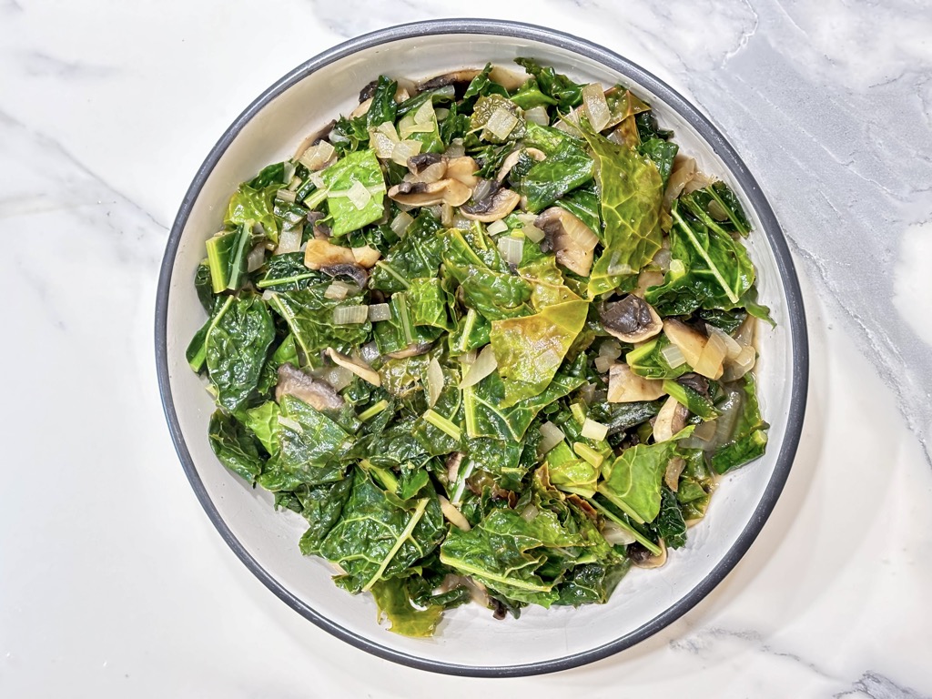 White dish filled with cooked kale and mushrooms on marble surface