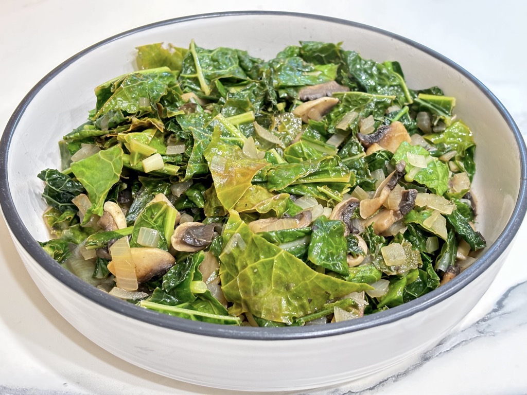 White dish filled with cooked kale and mushrooms on marble surface