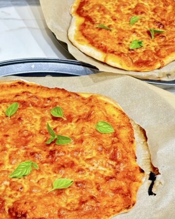 two margarita pizzas on baking sheets topped with basil