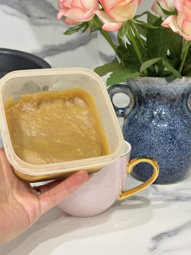 Hand holding small container of caramel sauce in front of pink and gold coffee cup, placed in front of blue vase of roses and coffee knock box