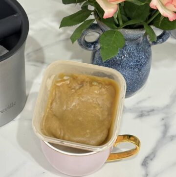 Small container of caramel sauce placed in pink and gold coffee cup, placed in front of blue vase of roses and coffee knock box