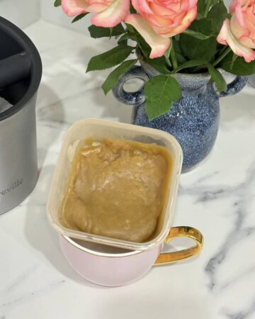 Small container of caramel sauce placed in pink and gold coffee cup, placed in front of blue vase of roses and coffee knock box