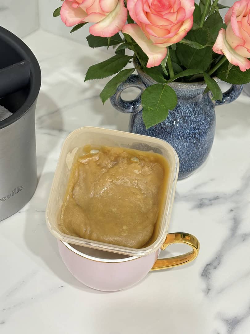 Small container of caramel sauce placed in pink and gold coffee cup, placed in front of blue vase of roses and coffee knock box