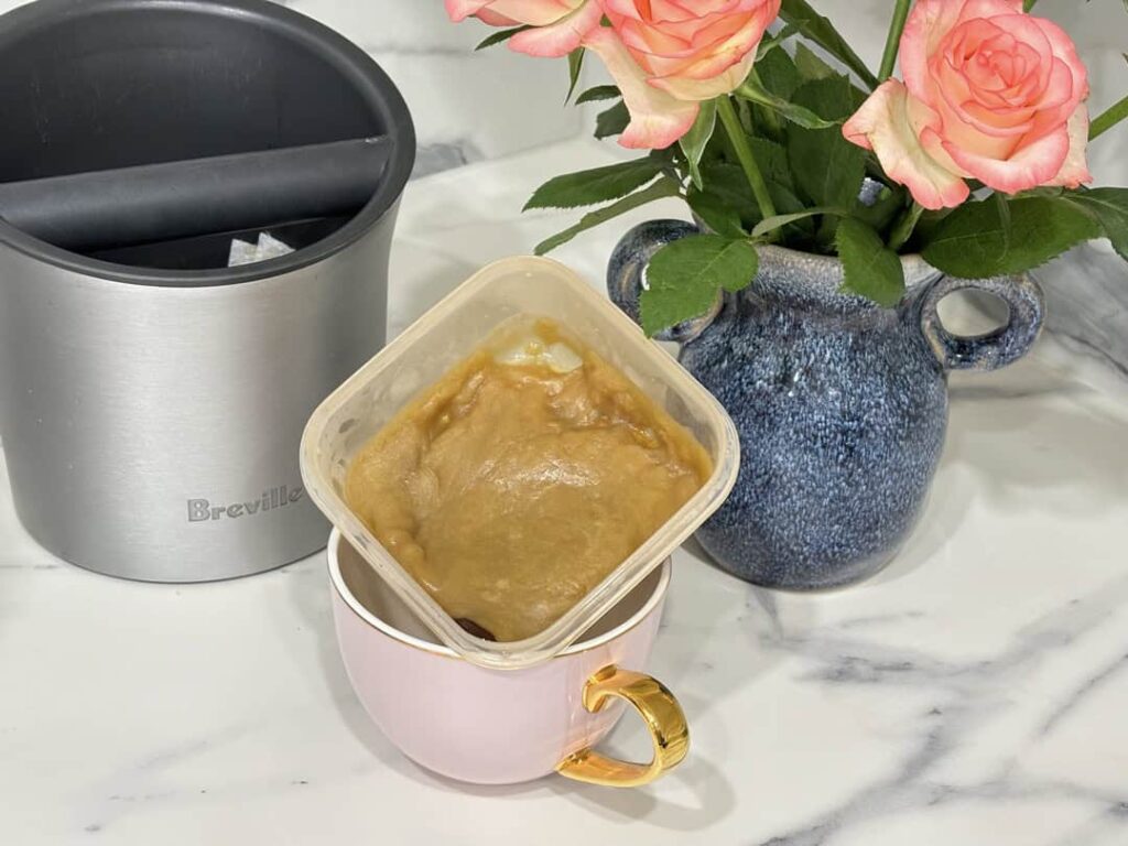 Small container of caramel sauce placed in pink and gold coffee cup, placed in front of blue vase of roses and coffee knock box