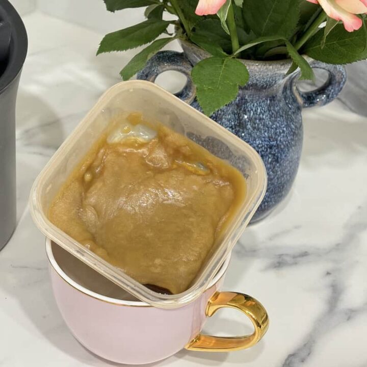 Small container of caramel sauce placed in pink and gold coffee cup, placed in front of blue vase of roses and coffee knock box