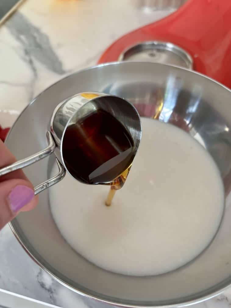 hand pouring measuring cup of maple syrup into silver bowl of coconut milk