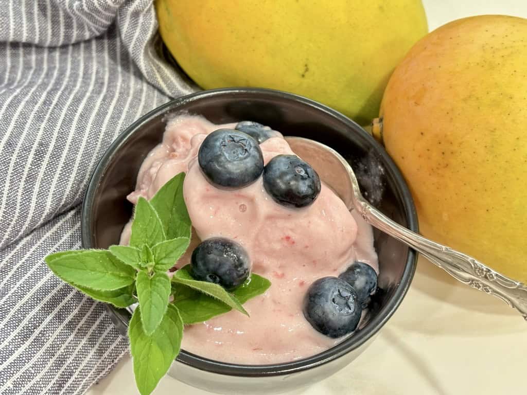 black bowl with silver spoon filled with raspberry ice cream topped with blueberries and herb in front of striped napkin and two ripe mangoes