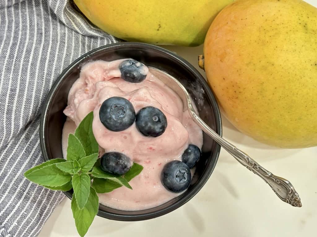 top down shot of black bowl with silver spoon filled with raspberry ice cream topped with blueberries and herb in front of striped napkin and two ripe mangoes