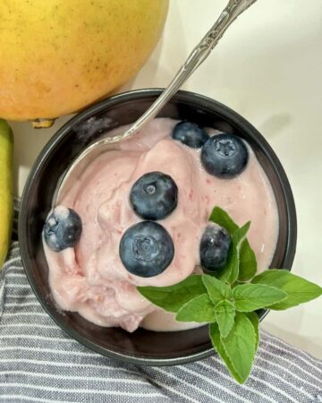 top down shot of black bowl with silver spoon filled with raspberry ice cream topped with blueberries and herb in front of striped napkin and two ripe mangoes