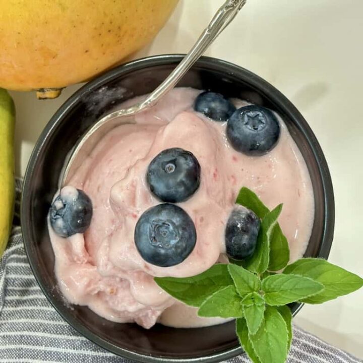 top down shot of black bowl with silver spoon filled with raspberry ice cream topped with blueberries and herb in front of striped napkin and two ripe mangoes