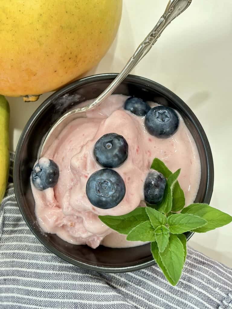 top down shot of black bowl with silver spoon filled with raspberry ice cream topped with blueberries and herb in front of striped napkin and two ripe mangoes